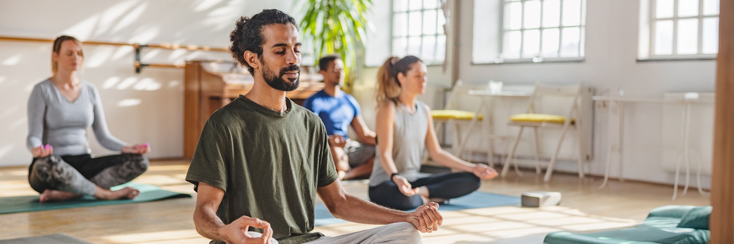 A group of people meditating in a yoga studio, with a man with dark hair in a pony tail in front.